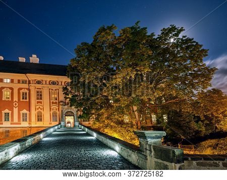 Stone Bridge Leading Towards A Historical Building At Night With The Moon Shining Through A Tree Cro