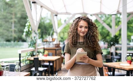 Waitress With Order Pad Standing Outdoors On Terrace Restaurant, Looking At Camera.