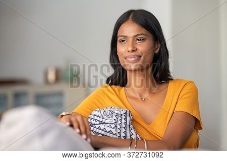 Young indian woman relaxing on couch at home while looking away. Thoughtful smiling mixed race girl daydreaming while sitting on sofa at home. Pensive middle eastern woman thinking.