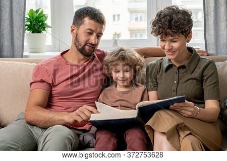 Happy young couple and their cute little son looking through pages of large encyclopaedia while sitting on comfortable couch by window