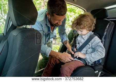 Young man helping his cute little son to put seatbelt while both going to countryside or supermarket on sunny summer day or morning