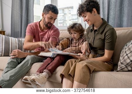 Young man and his little son pointing at page of open encyclopaedia while father explaining the subject of article or curious facts