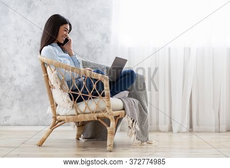 Young Asian Woman Working On Laptop And Talking On Cellhone While Sitting In Stylish Wicker Chair At