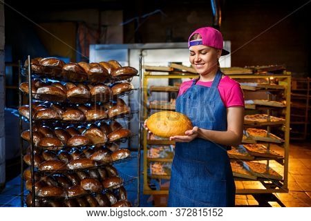 A Baker Girl Takes Hot Bread In A Bakery Against The Background Of Shelving With Bread. Industrial P