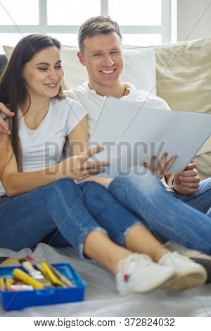 Young couple moving in to new home together, sitting on The floor and diskuss plan of desing.