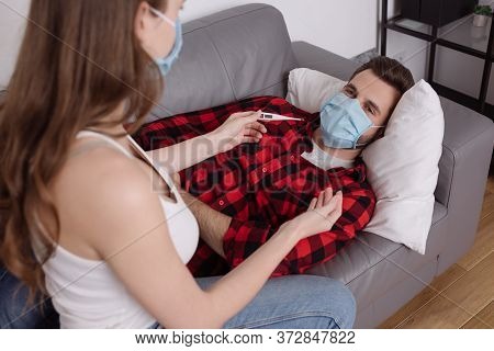 Focus Of Girl Holding Thermometer Near Diseased Boyfriend Lying On Sofa In Medical Mask