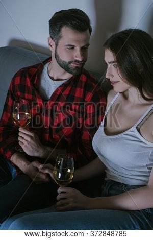Man Looking At Smiling, Dreamy Girl While Sitting On Sofa And Holding Glasses Of White Wine