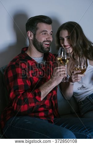 Young Couple Laughing While Sitting On Sofa And Holding Glasses Of White Wine