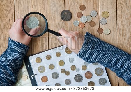 Woman-numismatist Views Coins From A Coin Album Through A Magnifying Glass. Top View