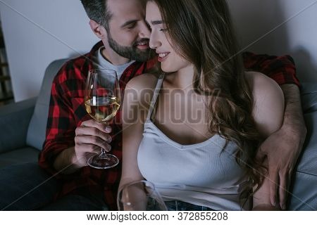 Man Embracing Happy Girlfriend While Sitting On Sofa With Glasses Of White Wine