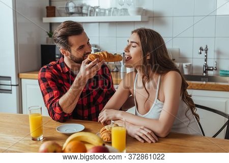 Man Feeding Pretty Girlfriend With Delicious Croissant Near Orange Juice And Fresh Fruits