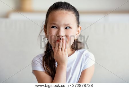 Chinese Girl Chuckling And Laughing Covering Mouth Sitting On Sofa At Home. Kids Laughter Concept.