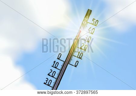 Thermometer Shows Heat In The Summer Season Against A Blue Sky With Clouds And Sun Rays, Weather Phe