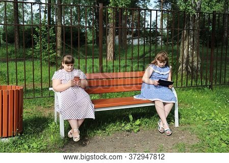 Chubby Young Girls Are Sitting On A Park Bench At A Social Distance. They Are Busy With Their Device