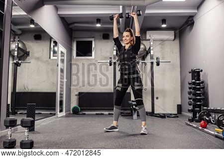 Electrical Muscle Stimulation. Woman In A Special Black Ems Suit Performs Squats And Dumbbell Arm Ex