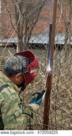 A Welder In A Camouflage Jacket And A Red Protective Mask With The Inscription In Russian 