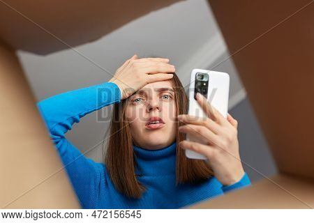 Shocked Disappointed Young Adult Woman Customer Wearing Blue Turtleneck Opening Cardboard Box, Looki