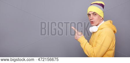 Side View Portrait Of Surprised Astonished Man In Beanie Hat And Yellow Casual Hoodie, Pointing Away