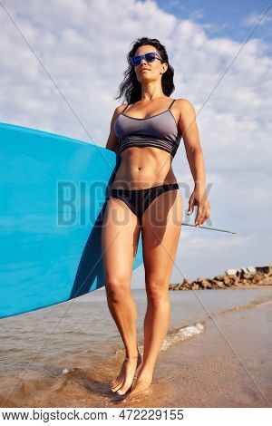 Charming Woman With Dark Hair And Athletic Tanned Body Smiling Carries A Windsurfboard In The Settin