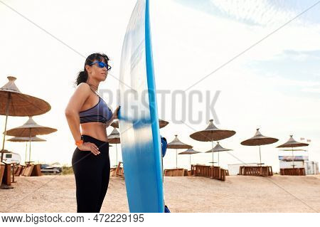 Charming Woman With Dark Hair And Athletic Tanned Body Smiling Carries A Windsurfboard In The Settin