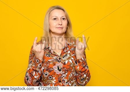 Caucasian Blonde Woman In A Patterned Dress Making The Rock Gesture With Both Hands, Isolated On Yel