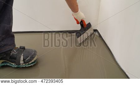 Repairman In Protective Gloves And Uniform Using A Metal Spatula To Level A Liquid Self-levelling Fl