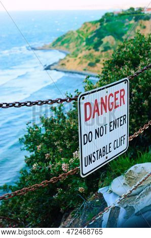 A Bold Sign, Attached To A Chain Link, Warns Visitors About The Danger Of An Unstable Cliff. Sandbag