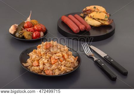 Stewed Cabbage - Bigos In A Black Plate Against The Background Of Pickled Vegetables And Baked Potat