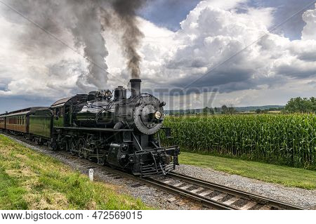 A Steam Passenger Train Passing Corn Fields And Blowing Smoke On A Sunny Summer Day
