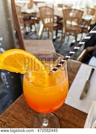An Orange Cocktail With A Straw And An Orange Slice In A Glass With A Soft Background Of A Restauran