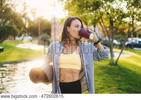 Beautiful Caucasian Woman Holding A Yoga Mat In The Park Drinking A Cup Of Tea Or Coffee And Looking