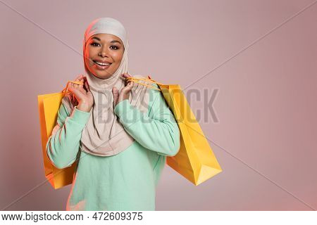 Overjoyed Multiracial Woman In Muslim Hijab Holding Yellow Shopping Bags And Smiling At Camera On Pi