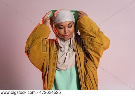 Young And Trendy Multiracial Woman In Hijab And Yellow Bomber Jacket Wearing Hood On Pinkish Grey Ba