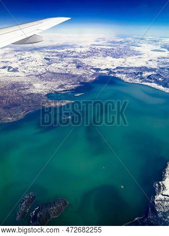 Aerial View Through Porthole Of Plane To Of Snow-covered Mountains And Mountain Lake. Scenic View Th
