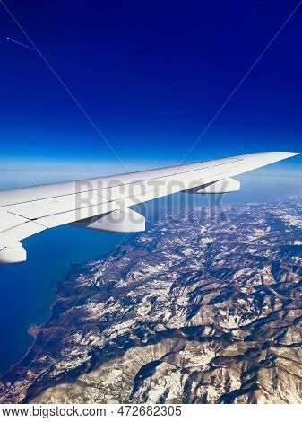 Aerial View Through Porthole Of Plane To Snow-covered Mountains And Sea. Scenic View Through Aircraf