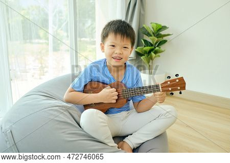 Cute Asian Happy Smiling 7 Years Old School Boy Sitting On Bean Bag Having Fun Playing Ukulele In Th