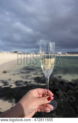 Hand With Glass Of White Champagne Or Cava Sparkling Wine On White Sandy Tropical Beach And Blue Oce