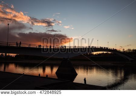 Silhouettes Of People On The Pedestrian Bridge In Novgorod Against The Sunset Sky With Pink Clouds