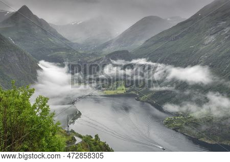 The Village Of Geiranger In Norway And The Fiord With Clouds And Fog And A Boat Sailing Over The Fio