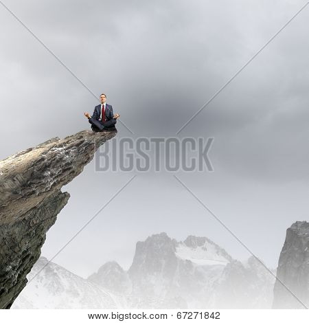 Young businessman sitting lotus pose on edge of rock mountain