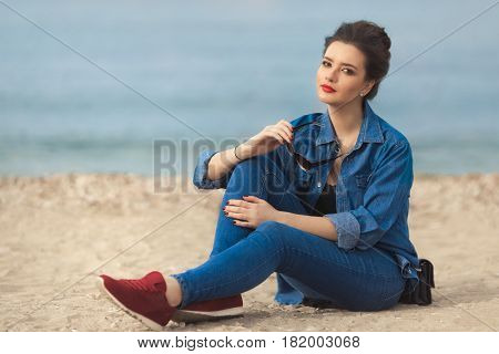 Denim outfit fashion details. Relaxing stylish woman enjoying the sun with red glitter manicure in navy jeans holding sunglasses and leather small cross body bag sitting on the beach.