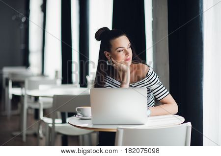 Portrait of an attractive woman sitting in some coffee shop with her laptop and thinking over a subject while looking through the window