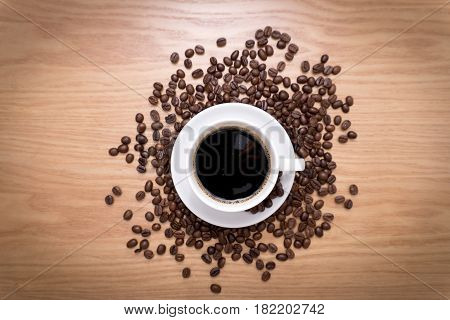 White cup with black, hot coffee standing on brown, roasted beans on wood table, close up.