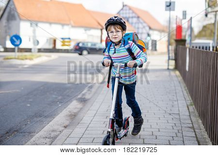 Active school kid boy in safety helmet riding with his scooter in the city with backpack on sunny day. Happy child in colorful clothes biking on way to school. Safe way for kids outdoors to school.