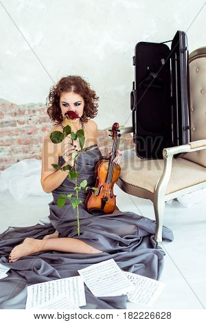 Front view beautiful woman sitting on the floor near chair and holding a violin and smelling red rose flower in studio