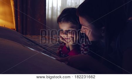 Close-up of mother and her little daughter watching tablet. Cute girl looking at device while chilling at home.