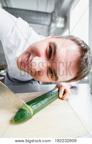 Young male chef in uniform cutting cucumber and looking crazily at camera while having fun.