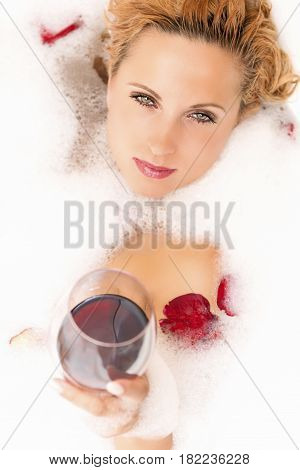 Closeup of Passionate Relaxing Sensual Caucasian Blond Female in Foamy Bathtub. Holding a Glass of Red Wine. Vertical Image Orientation