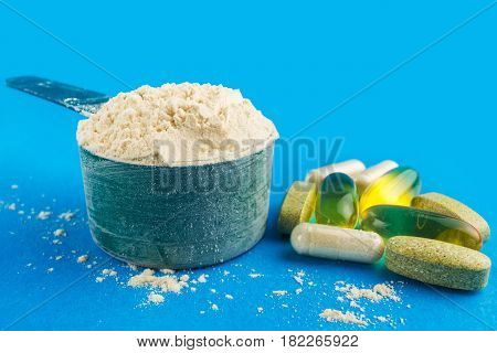 Protein powder in measuring plastic spoon and vitamin complex omega 3 glucosamine capsules supplements on blue background sports nutrition