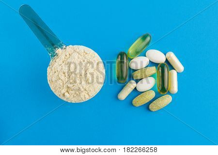 Protein powder in measuring plastic spoon and vitamin complex omega 3 glucosamine capsules supplements on blue background sports nutrition top view.